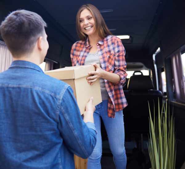 Portrait of young happy woman unloading cardboard boxes from moving van handing them to husband